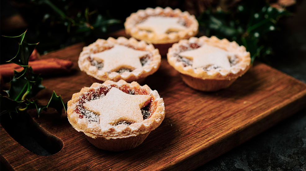 Four Mince pies dusted with icing sugar on a dark wood board with Holly and Pine cones.